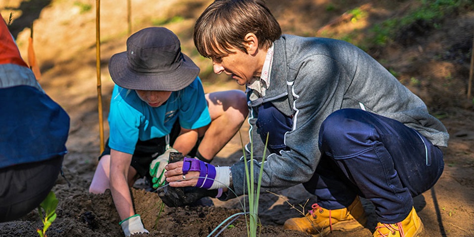 Woman and primary school student crouched on the ground planting tubestock