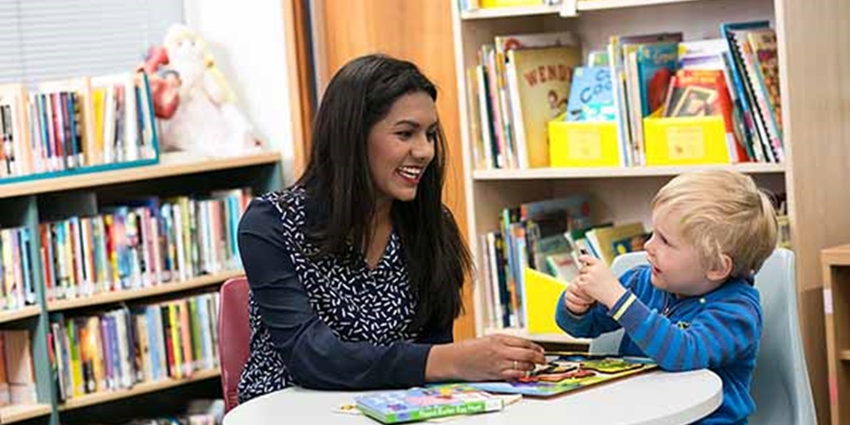 Teacher sits with early childhood student in library.