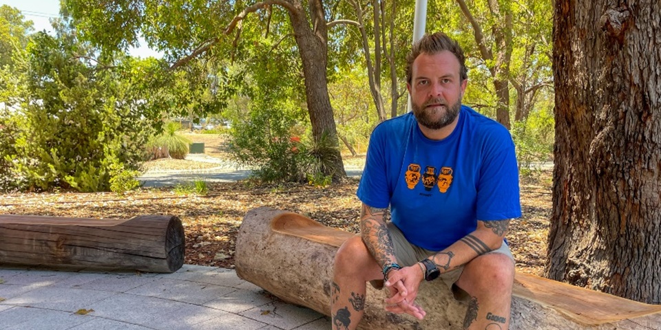 Man sitting on tree log in bush setting looking towards camera